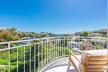 A sunny balcony with a wooden chair overlooks a lush, green hillside town and the blue sea under a clear sky.