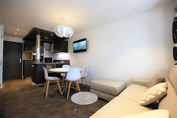 A sleek, modern open-concept living space features a dark kitchen, white dining set, light sofa, ottoman, and wall-mounted TV.