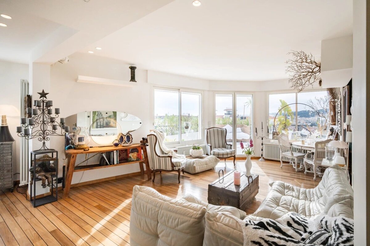 A bright, spacious living room with wooden floors, white sofas, eclectic decor, and large windows leading to a balcony.