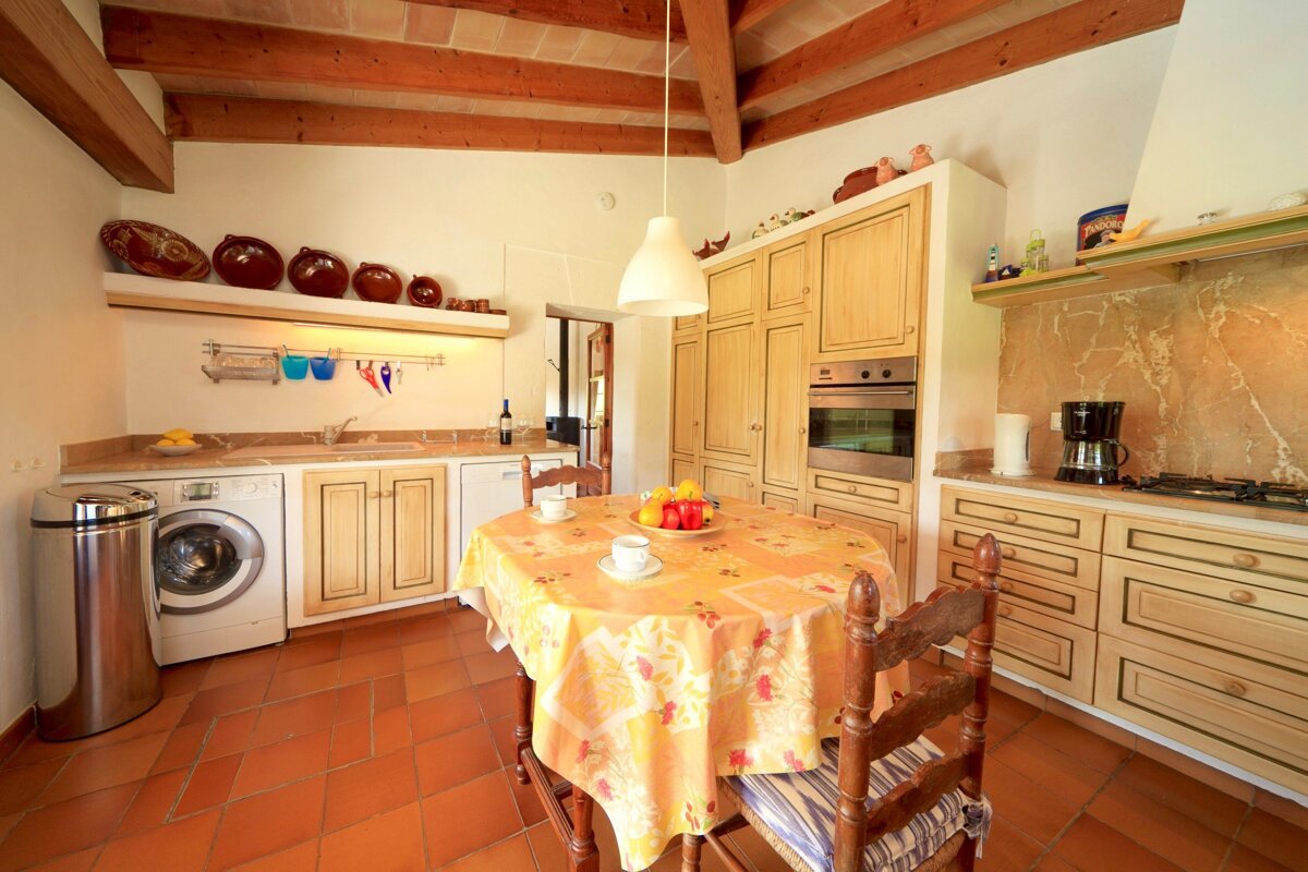Warm, rustic kitchen with terracotta floor, wooden beams, and light wood cabinets. A central table, appliances like a washer and oven, and traditional decor fill the room.