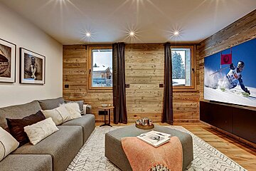 A modern, rustic living room featuring wood-paneled walls, a large grey sofa, and windows with snowy views. A big TV shows a ski race, creating a cozy chalet vibe.