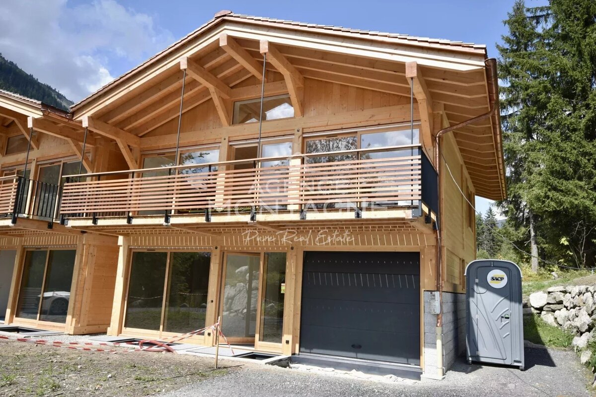 A large, light-wood chalet with multiple balconies, large windows, a garage door, and a portable toilet next to it, against a backdrop of trees and mountains.
