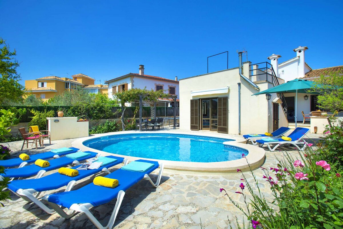 Sunny pool area with blue loungers, a white villa, and lush gardens under a clear sky. Perfect for a relaxing vacation.