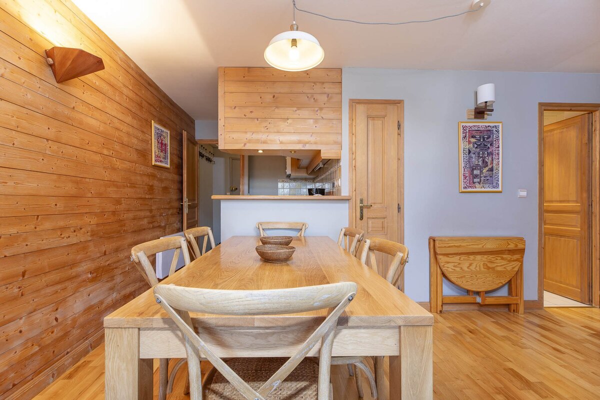 A cozy, rustic dining area with a long wooden table and chairs, featuring wood-paneled walls, a light blue wall, and an open kitchen view.