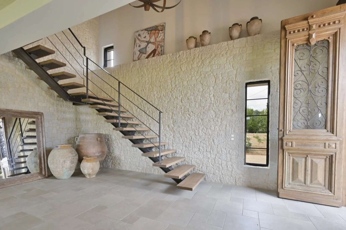 A modern floating wooden staircase ascends against a textured stone wall in a rustic interior. Large antique pots, an ornate wooden door, and a mirror complete the unique space.
