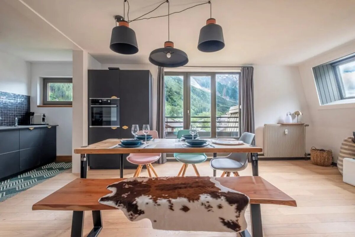 Modern dining room featuring a wooden table, colorful chairs, a cowhide-draped bench, pendant lights, and scenic mountain views.