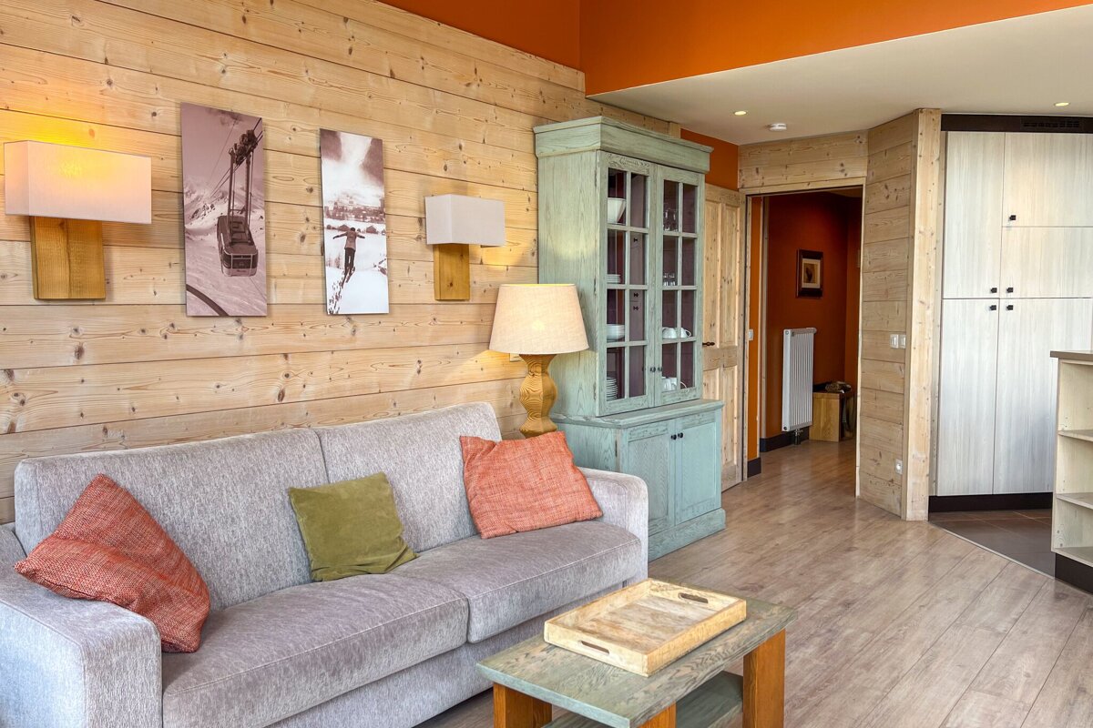 A cozy, wood-paneled living room with a gray sofa, orange ceiling, mint cabinet, ski-themed art, and warm lighting.