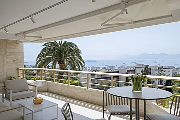 A modern luxury balcony with outdoor seating overlooks a palm tree and a scenic ocean view with boats and distant hills.