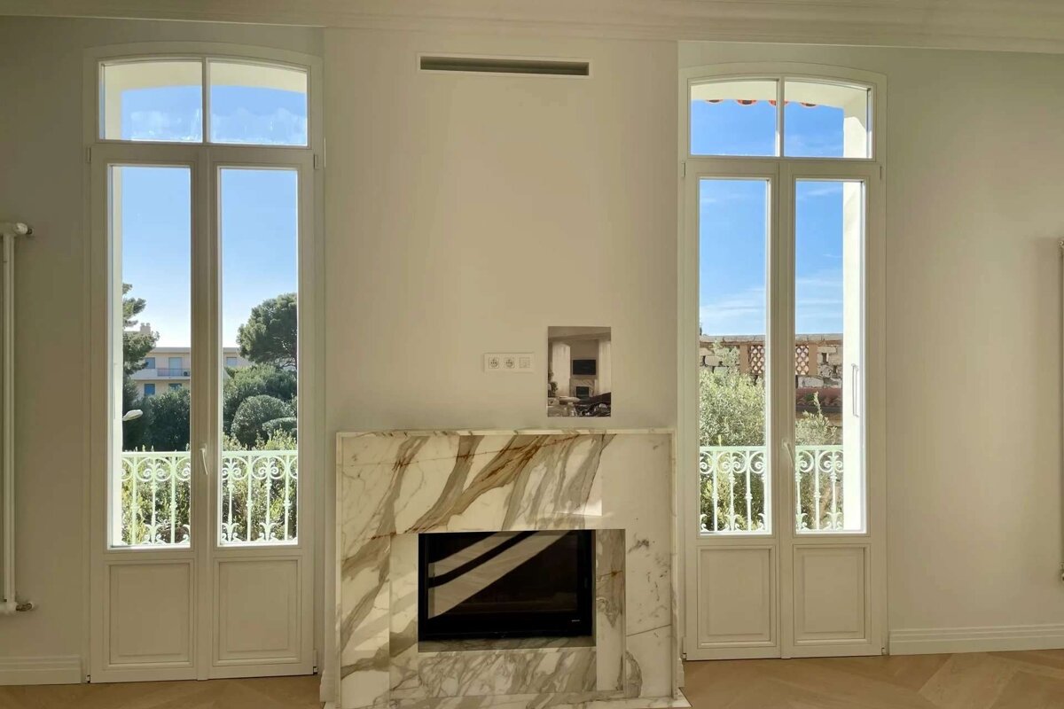 A light-filled room features two arched windows framing a green outdoor view, separated by a marble fireplace and a wall with a small recessed area.