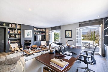 A spacious room featuring a study desk, lounge area with bookshelves, and open doors leading to a patio with scenic mountain views.