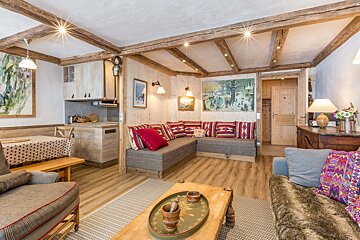 Cozy chalet living space with rustic wood beams, paneling, and a comfy L-shaped sofa. Features a dining nook and compact kitchen.