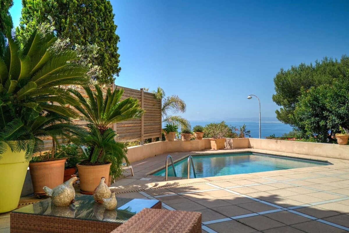 A sunny outdoor pool area overlooking the blue ocean, surrounded by lush potted palms and green trees, with a tiled patio and garden decor.