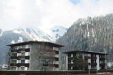 Two multi-story buildings with balconies stand before snow-covered mountains and dense pine forests under a cloudy sky, suggesting an alpine setting.