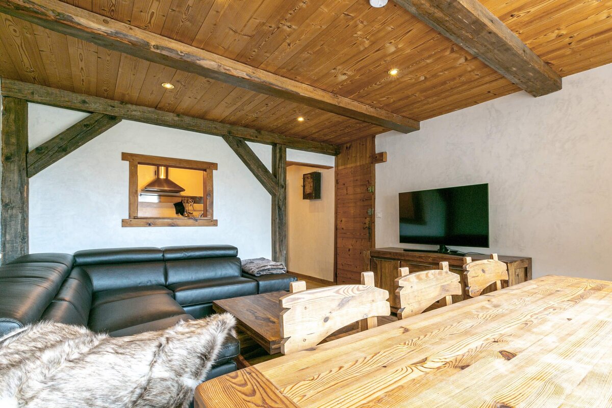 Chalet-style living/dining room with wood ceilings/beams, black leather sofa, rustic wooden table and chairs, and a TV. Kitchen seen through pass-through.
