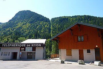 A green-treed mountain with a visible cable car line, behind the Téléphérique de Nyon station and a wooden chalet on a clear day.