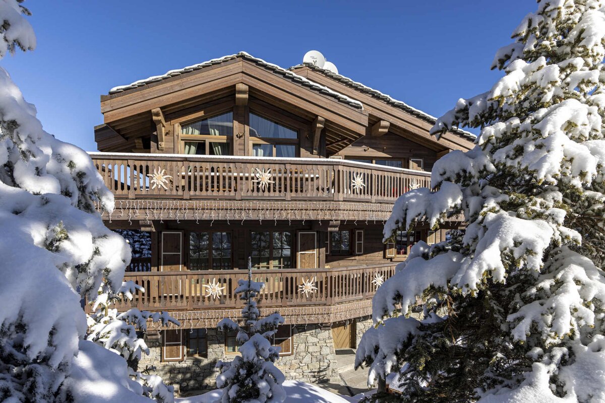 A large wooden alpine chalet framed by snow-covered pine trees under a clear blue winter sky.