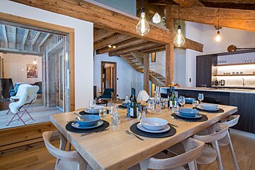 Rustic-modern open-plan living space featuring a large wooden dining table set for dinner, exposed beams, a stylish kitchen, and a cozy room beyond.