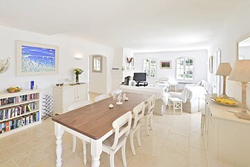 A spacious, bright white living/dining room with a wooden dining table, white furniture, a fireplace, bookshelves, and large windows.