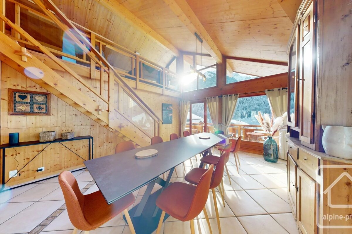 Cozy wooden chalet dining room featuring a long table, orange chairs, staircase to a loft, and sunny mountain views.