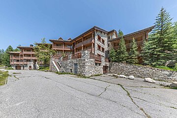 A large, multi-story stone and wood building with balconies, surrounded by pine trees, next to an cracked asphalt road under a clear sky.