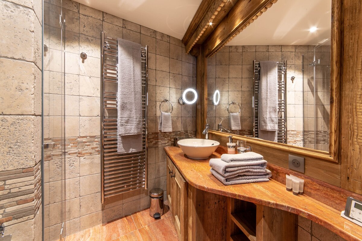 A warm, rustic bathroom featuring wood and stone tiles, a vessel sink, heated towel rail, and a large mirror with wooden frame.