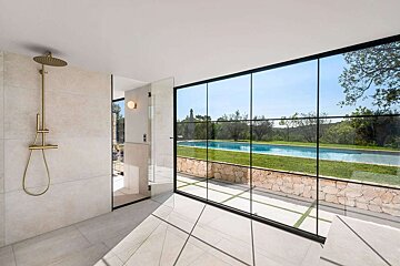 A modern bathroom features a brass shower and floor-to-ceiling windows, offering a scenic view of a swimming pool, green landscape, and blue sky.