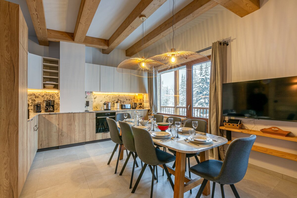 Modern kitchen/dining room with exposed wooden beams, a set table for six, appliances, and a large window revealing a snowy winter landscape.