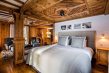 A cozy bedroom featuring extensive wood paneling on walls, floor, and a patterned ceiling, a modern bed, armchair seating, and mountain art.