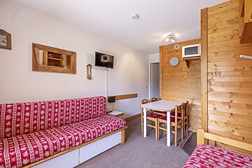A small living area featuring a red patterned sofa, a white dining table with chairs, a wall-mounted TV, and a microwave on a wood-paneled wall.
