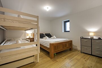 Bright bedroom with white bunk beds, a wooden double bed, and a dresser on light wood flooring. A small window adds natural light.