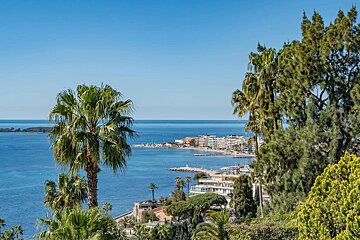 Scenic coastal view of a resort town along the blue Mediterranean Sea, framed by palm trees and lush greenery under a clear sky.