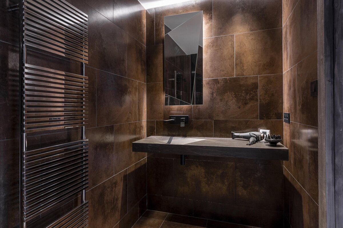 Modern dark brown tiled bathroom with a backlit, geometric mirror, floating sink, chrome towel warmer, and a hairdryer on the counter.