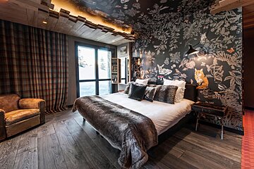 Rustic bedroom with a dark, woodland-patterned wall, a large bed, faux fur blanket, wooden floors, and a window revealing a snowy scene. Cozy chalet vibe.