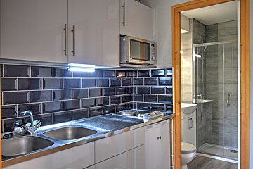 A compact kitchenette with white cabinets, black subway tiles, sink, stove, and microwave. A mirror reflects a small bathroom with a shower and toilet.