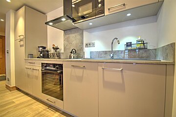 A sleek, modern kitchen with light beige cabinets, grey counters, and wood-effect flooring. It features a sink, oven, microwave, and appliances.