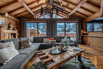 A cozy, rustic wooden chalet living room with a gray sectional, unique coffee table, and large windows revealing a beautiful snowy mountain view.