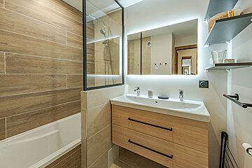 A modern bathroom featuring wood-patterned tiles, a white bathtub, a light wood vanity, and a large backlit rectangular mirror.