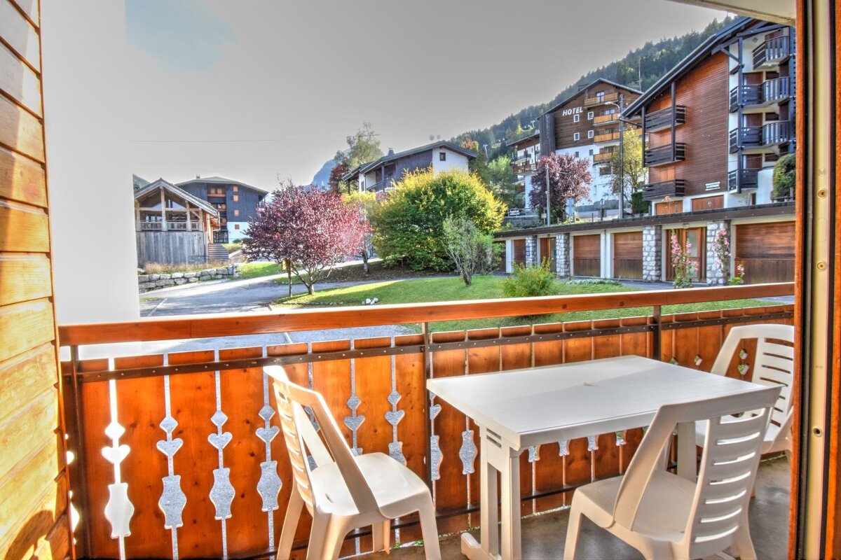 A balcony with white furniture overlooks a sunny mountain village featuring chalet-style buildings, garages, and colorful trees.