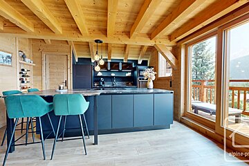 A cozy, modern chalet kitchen features light wooden walls/beams, dark blue cabinets, a central island with teal bar stools, and large windows revealing a mountain view.