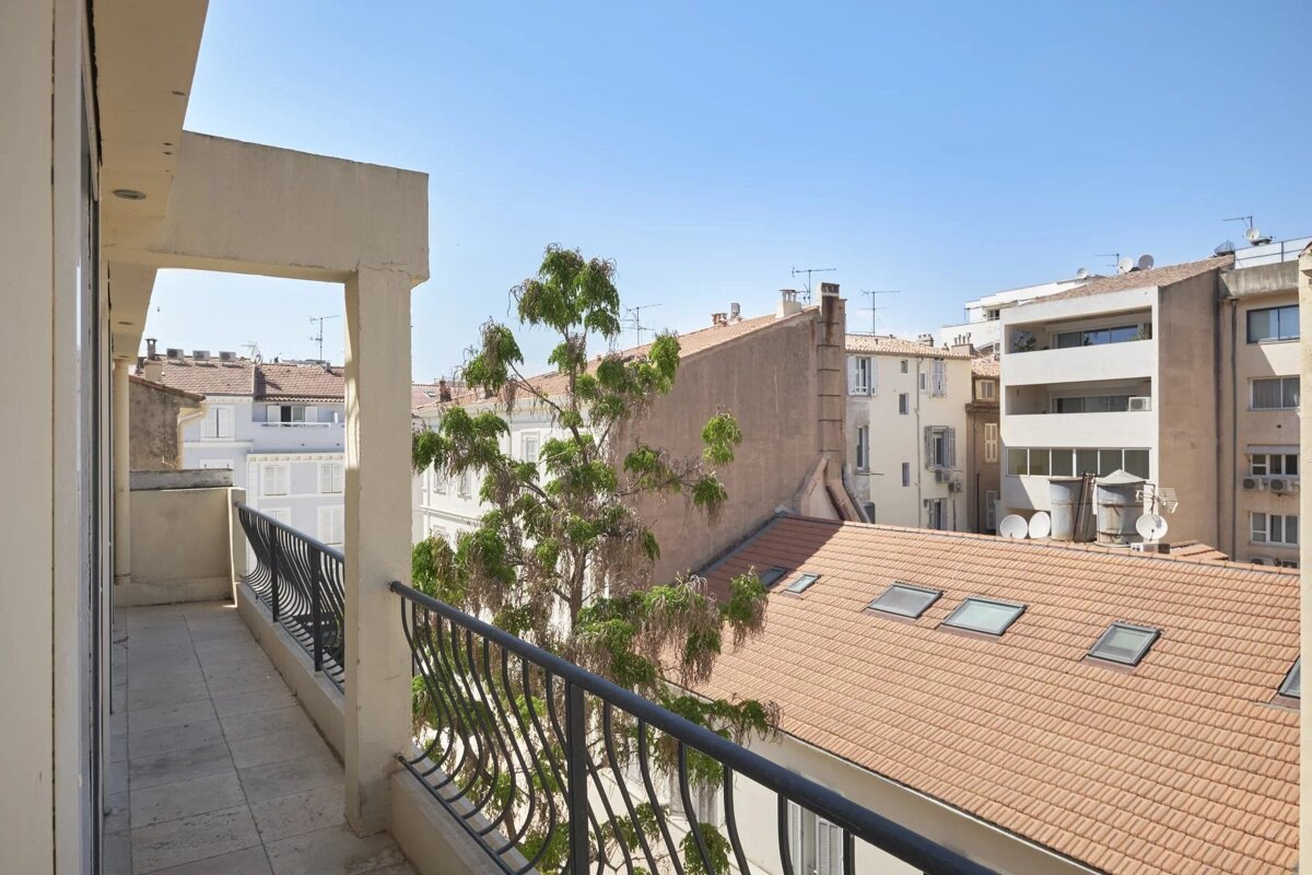 Balcony view over a city with buildings, terracotta roofs, a tree, and a clear blue sky on a sunny day.