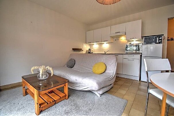 A studio apartment with a futon sofa, a wooden coffee table, and a small kitchenette with white cabinets.
