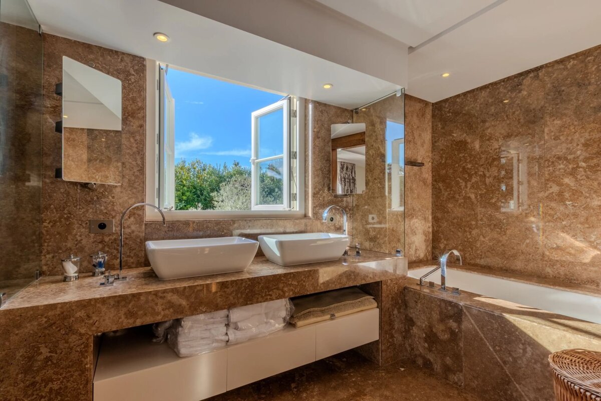 Luxurious brown marble bathroom with dual sinks, a tub, and a large open window showing green trees and blue sky.