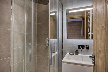 Modern bathroom featuring a glass shower cabin, grey tiled walls, a white vanity sink, and a backlit mirror.