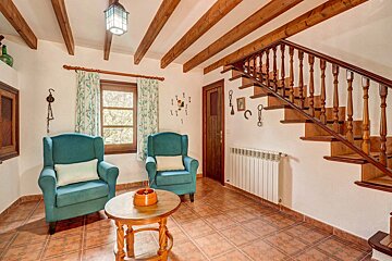 A cozy, rustic living room featuring teal armchairs, a grand wooden staircase, exposed ceiling beams, and tiled floors. A window with floral curtains brightens the space.