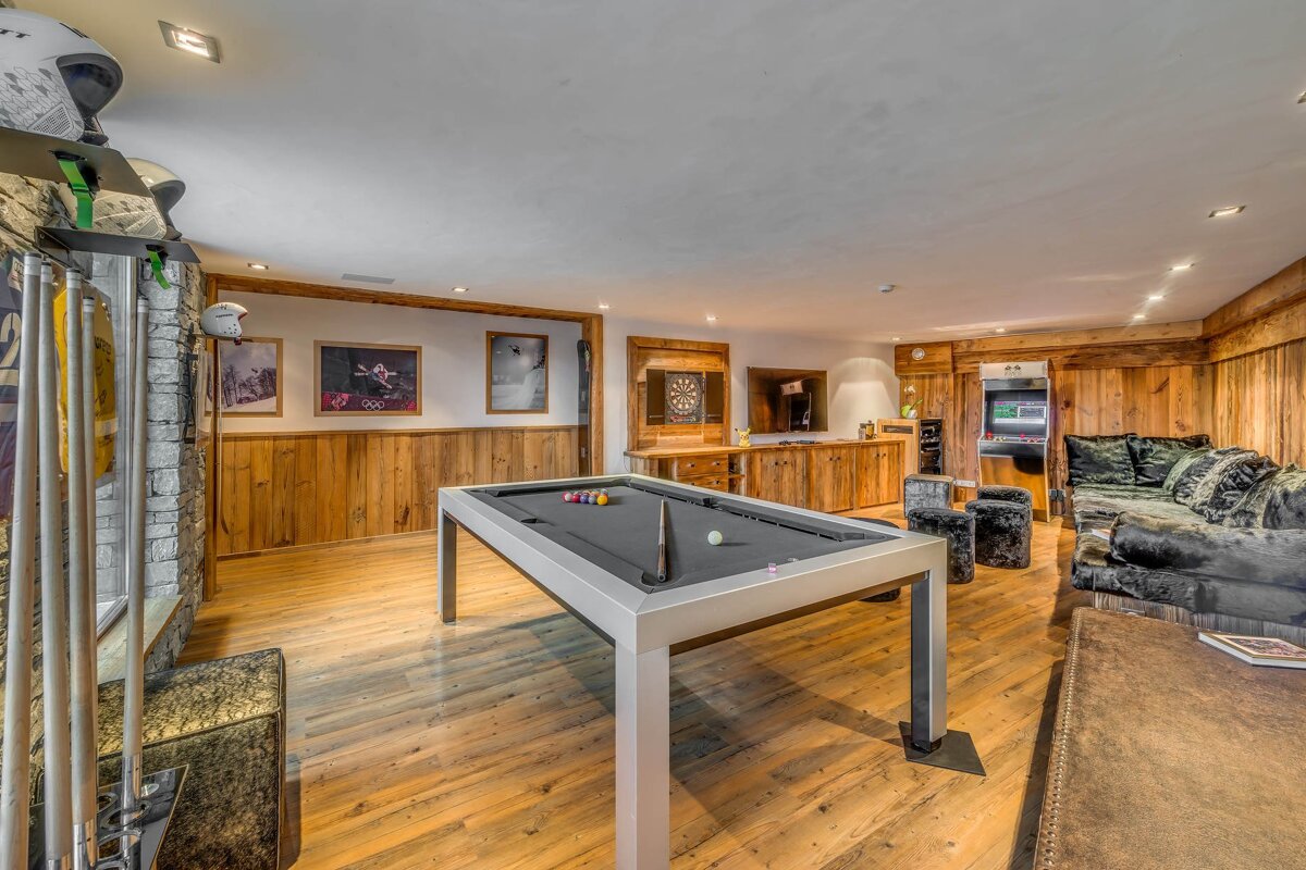 A cozy wooden game room featuring a modern grey pool table, plush seating, ski decor, and an arcade cabinet.
