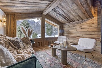 Cozy, rustic wood-paneled chalet living room with plush furnishings, warm lighting, and large windows overlooking a snowy mountain scene.