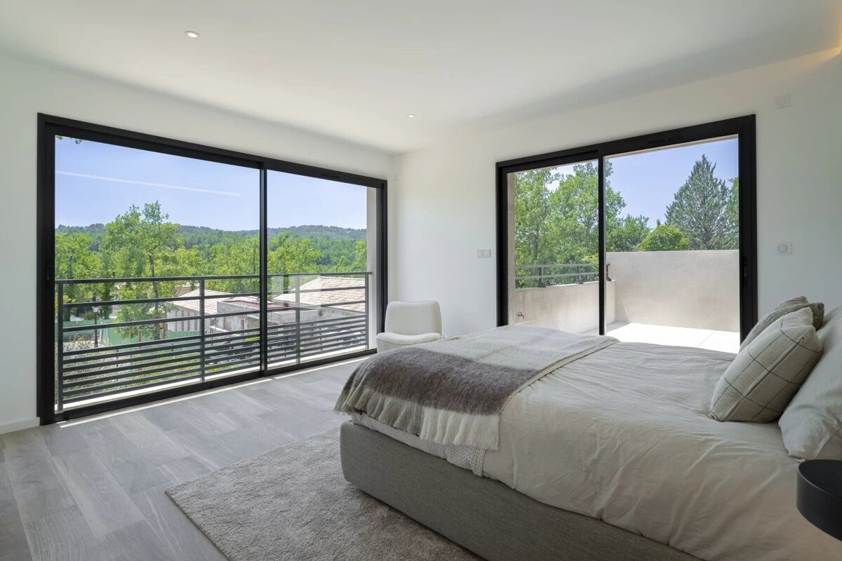 A bright, modern bedroom with large windows offering a panoramic view of a lush green forest, featuring a bed and a small balcony.