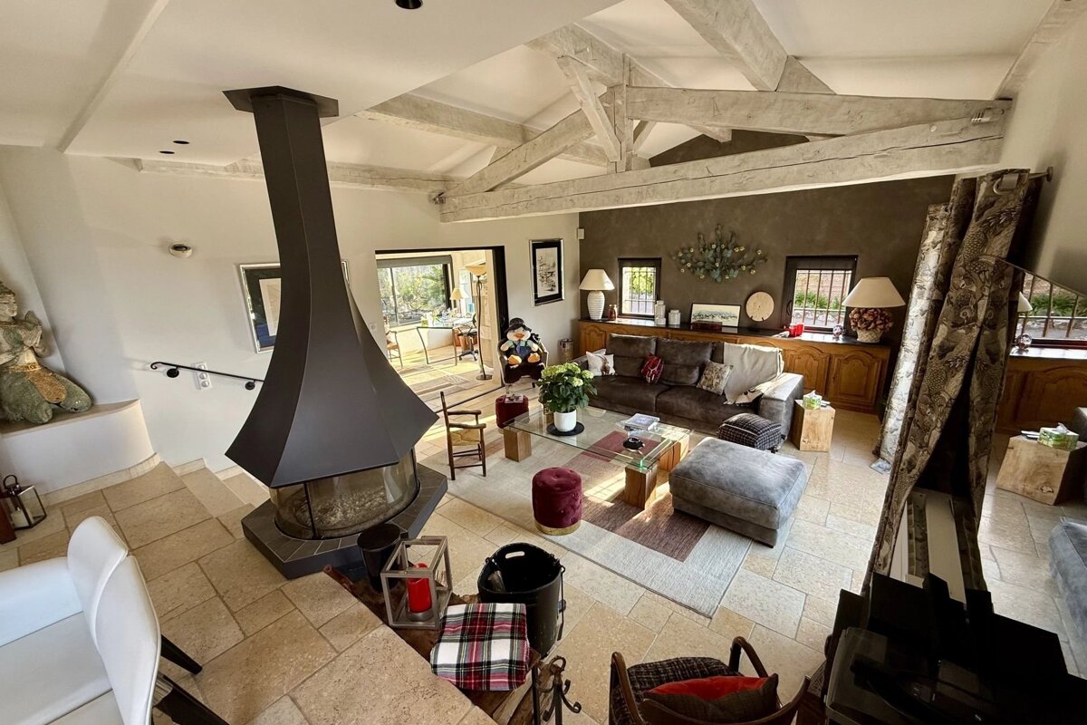 A cozy, rustic living room with exposed ceiling beams, a large central black fireplace, stone tile floors, and comfortable seating.