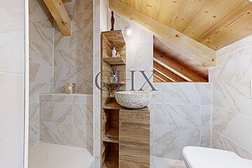 Rustic attic bathroom featuring sloped wooden ceilings, marble-patterned tiles, and a wooden vanity with a stone vessel sink.