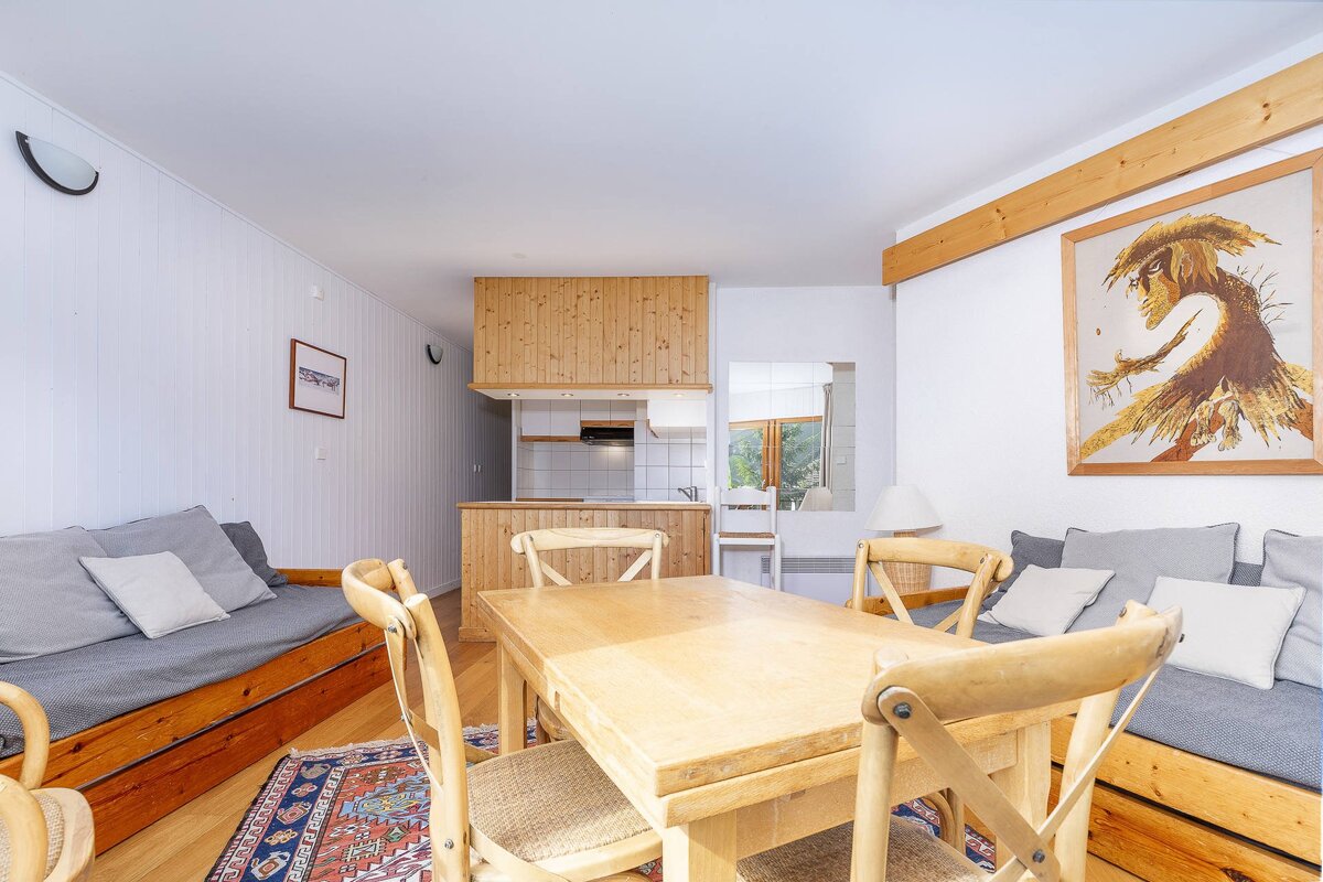 Rustic open-plan living/dining room with a wooden kitchen. Features a large wood table, built-in sofas, white walls, wood accents, and an ornate rug.